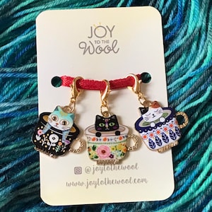 May include: Three gold cat-themed stitch markers with red string. Each marker features a cat in a teacup with a different colored cat and teacup design. The text "JOY TO THE Wool" is on the white card behind the markers.