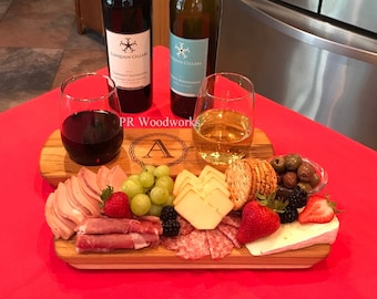 May include: A wooden serving board with a personalized engraved letter "A" and the text "PR Woodworks". The board is filled with a charcuterie assortment including cheese, grapes, crackers, olives, salami, prosciutto, and berries. Two glasses of wine and two bottles of wine are in the background.