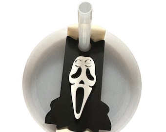 May include: A white tumbler with a clear straw and a black and silver Scream mask charm. The charm has a cutout design of the mask's face. The text "CRISTY COLOMBO" is printed on the charm.