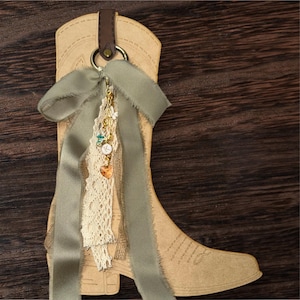 May include: A tan cowboy boot-shaped tag with a brown leather strap and a gold ring. A sage green ribbon and cream lace are attached, adorned with charms including a heart, butterfly, and clock. The background is a dark wood grain.