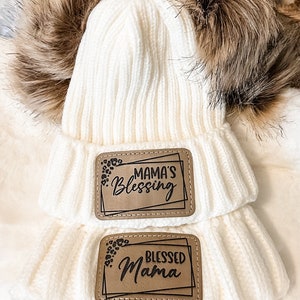 May include: White knitted beanie with two brown faux fur pom-poms. The beanie has two brown leather patches with black text. The top patch says "Mama's Blessing" and the bottom patch says "Blessed Mama".