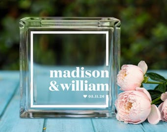 May include: A clear glass block with the names "madison & william" and the date "05.11.26" in white lettering. The block is decorated with a white border and sits on a light blue surface, with pink flowers in the foreground.