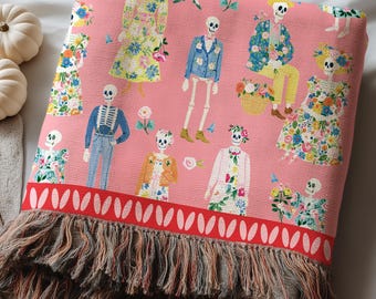 May include: A pink woven throw blanket featuring a repeating pattern of skeletons dressed in colorful floral outfits and casual clothing. The blanket has a wide red border with a white oval design and a long, multi-colored fringed edge at the bottom.