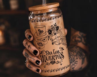 May include: A clear glass can-shaped tumbler filled with a light brown iced beverage, featuring a black graphic print of two sugar skulls and the text "DIA LOS MUERTOS". The glass has a bamboo lid and a clear glass straw inserted through the top.