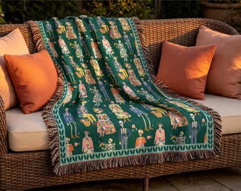 May include: A dark green woven throw blanket featuring a repeating pattern of skeletal figures dressed in colorful floral clothing and suits, finished with a decorative leaf-patterned border and fringed edges on the top and bottom sides.