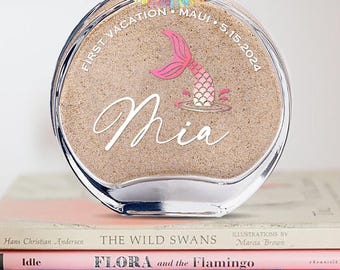 May include: A clear glass bottle filled with sand, featuring a mermaid tail design and the name "Mia." The bottle is adorned with a colorful tassel and a cork stopper. Text on the bottle reads "First Vacation Maui 5.15.2024."
