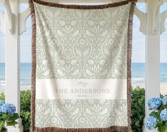May include: A woven throw blanket featuring a sage green and cream botanical pattern with birds and flowers. A horizontal cream band across the center displays the text THE ANDERSONS and the date 06.15.26. The blanket has a multicolored fringe.