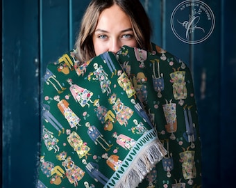 May include: A dark green woven throw blanket featuring a repeating pattern of skeletons dressed in colorful floral clothing. The blanket has a decorative border with a leaf motif and light-colored fringe along the bottom edge of the fabric.