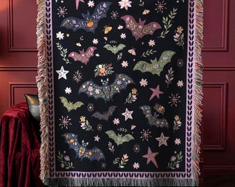 May include: A black woven throw blanket featuring a repeating pattern of bats, stars, and floral motifs in shades of pink, green, and orange. The blanket has a decorative border with a leaf pattern and frayed edges along the top and bottom sides.