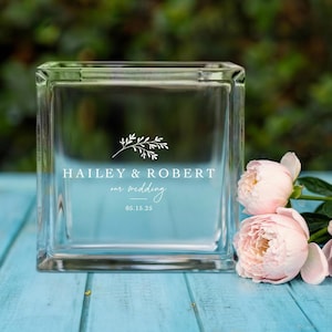 May include: A clear glass block with the text "HAILEY & ROBERT our wedding 05.15.25" in white. The block is sitting on a light blue surface with pink flowers in the background.