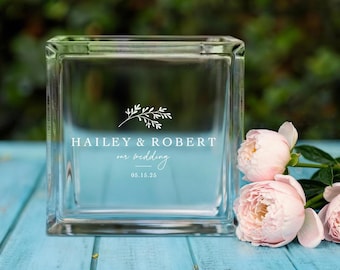 May include: A clear glass block with the text "HAILEY & ROBERT our wedding 05.15.25" in white. The block is sitting on a light blue surface with pink flowers in the background.