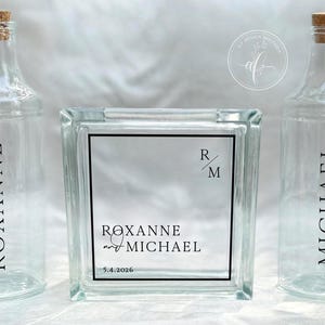 May include: Clear glass bottles with cork stoppers and a glass block with the names "Roxanne" and "Michael" printed on them. The block also has the date "5.4.2026" printed on it. The bottles and block are likely for a wedding or event.