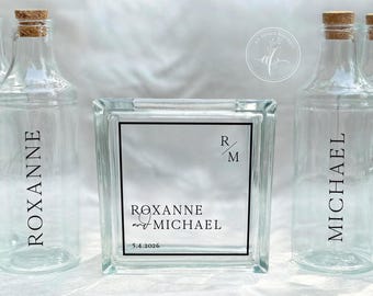 May include: Clear glass bottles with cork stoppers and a glass block with the names "Roxanne" and "Michael" printed on them. The block also has the date "5.4.2026" printed on it. The bottles and block are likely for a wedding or event.