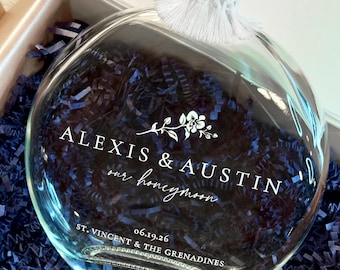 May include: A round clear glass bottle with a cork stopper and white tassel trim. The front features white text reading ALEXIS & AUSTIN, our honeymoon, 06.19.26, and ST. VINCENT & THE GRENADINES, accompanied by a small floral graphic illustration.