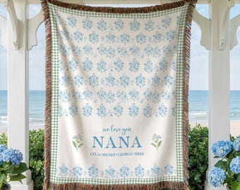 May include: A woven throw blanket featuring a light blue floral pattern on a cream background with a green and white gingham border. The text reads "we love you NANA" followed by the names "LYLA • HENRY • GEORGE • BREE" in blue lettering.