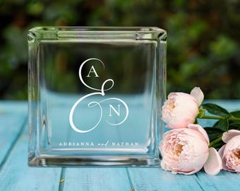 May include: A clear glass block with the initials "A & N" in white script, and the names "Adrianna and Nathan" below. Pink peonies rest on a blue wooden surface, with a blurred green background.