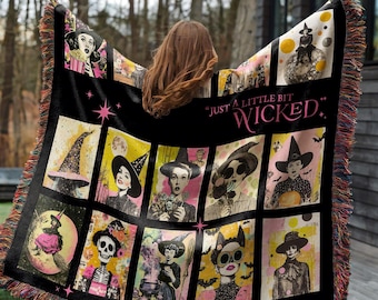 May include: A black woven throw blanket featuring a grid of illustrations depicting witches, skeletons, and magical elements in yellow, pink, and black. The text "JUST A LITTLE BIT WICKED" is printed in pink. The blanket has a multi-colored fringed edge.