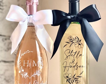 May include: Two wine bottles, one rose and one white, are decorated for a wedding. The rose bottle has a white bow and the initials "HM" with "Hanna & Michael" written below. The white wine bottle has a black bow and the names "Stella & Hudson" with a date.