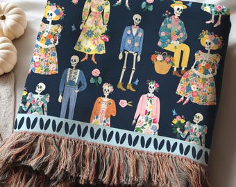 May include: A dark navy blue woven throw blanket featuring a repeating pattern of skeletons dressed in colorful floral outfits and suits. The blanket has a light blue border with a dark leaf print and a long, multi-colored fringed edge at the bottom.