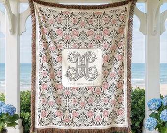 May include: A woven throw blanket featuring a repeating floral pattern of pink flowers and dark green foliage on a cream background. A central white rectangular patch displays a large, ornate gray letter H and the text THE HAWTHORNS.