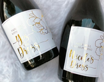 May include: Two bottles of sparkling wine with gold foil labels. The labels have floral designs and the text "Pairs well with saying Yes to the dress" and "Pairs well with finding Nicole's dress".