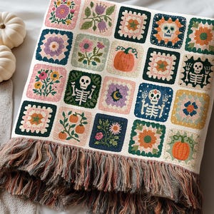 May include: A woven throw blanket featuring a grid of square motifs with various designs including skeletons, pumpkins, flowers, and celestial elements. The blanket has a cream-colored base and a multi-colored fringed edge in shades of brown and orange.