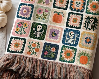 May include: A woven throw blanket featuring a grid of square motifs with various designs including skeletons, pumpkins, flowers, and celestial elements. The blanket has a cream-colored base and a multi-colored fringed edge in shades of brown and orange.
