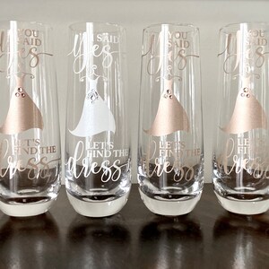Pu&ograve; includere: Quattro flute da champagne con il testo "You said yes, let's find the dress" in lettere dorate. Le flute sono decorate con una silhouette di abito bianco e dorato.
