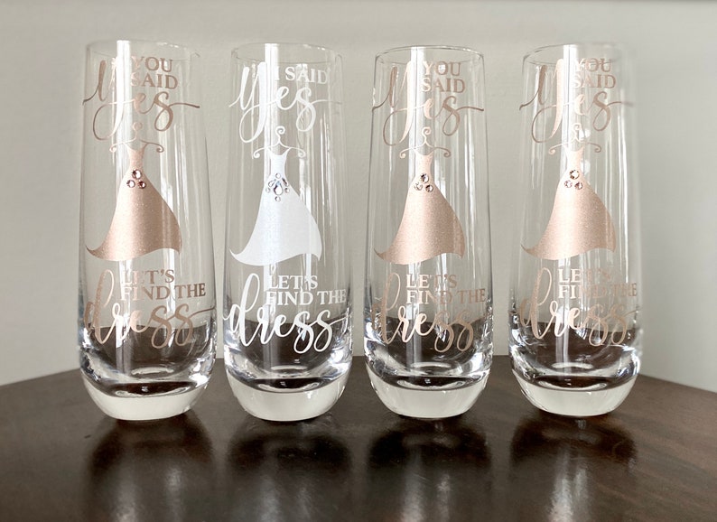 Pu&ograve; includere: Quattro flute da champagne con il testo "You said yes, let's find the dress" in lettere dorate. Le flute sono decorate con una silhouette di abito bianco e dorato.