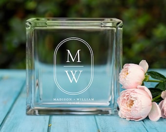 May include: A clear glass block with a white oval design featuring the letters "M" over "W". Below the letters is the text "MADISON + WILLIAM 05.11.16". The block is on a light blue surface with pink flowers.