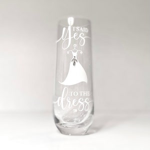 Pu&ograve; includere: Un flauto da champagne in vetro trasparente con il testo "It said yes to the dress" inciso sul lato. Il testo &egrave; in un font corsivo bianco e include un disegno di un abito da sposa.