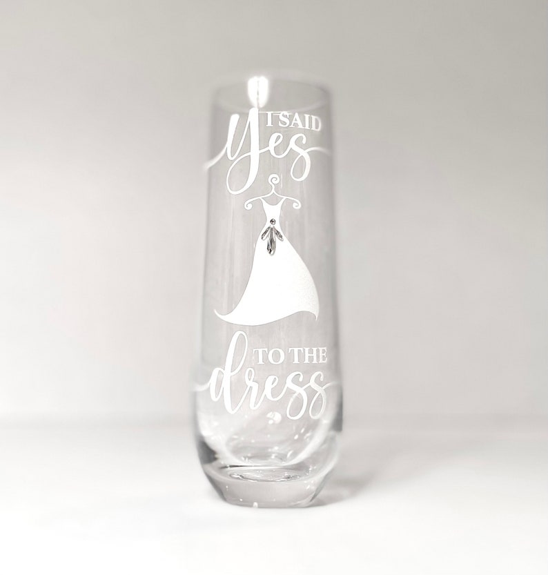 Pu&ograve; includere: Un flauto da champagne in vetro trasparente con il testo "It said yes to the dress" inciso sul lato. Il testo &egrave; in un font corsivo bianco e include un disegno di un abito da sposa.