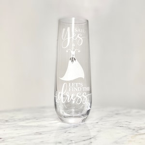 Pu&ograve; includere: Un flauto da champagne in vetro trasparente con il testo "Said Yes Let's Find the Dress" e una silhouette bianca di un abito da sposa.
