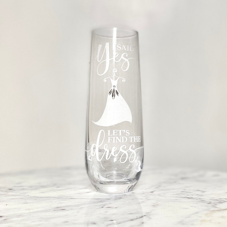Pu&ograve; includere: Un flauto da champagne in vetro trasparente con il testo "Said Yes Let's Find the Dress" e una silhouette bianca di un abito da sposa.