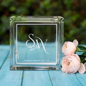 May include: A clear glass block with a white border and a light blue background. The block features the initials "S" and "W" in a decorative font, with floral accents. The text "SOPHIA + WESLEY | 05.11.26" is also visible.