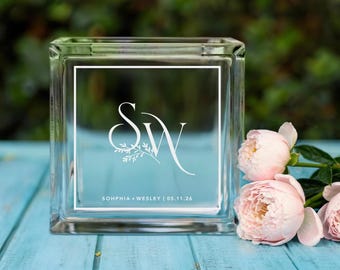 May include: A clear glass block with a white border and a light blue background. The block features the initials "S" and "W" in a decorative font, with floral accents. The text "SOPHIA + WESLEY | 05.11.26" is also visible.