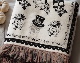 May include: A cream-colored woven throw blanket featuring a repeating black graphic print of sugar skulls adorned with flowers, top hats, and bow ties. The blanket has a decorative scalloped border and a multi-colored fringed edge at the bottom.