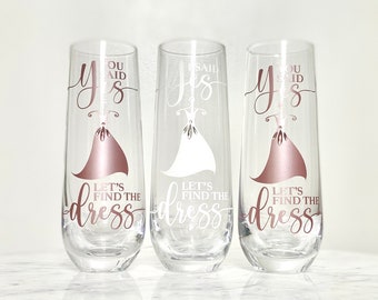 May include: Three champagne flutes with the text "You said yes, let's find the dress" on them. The flutes are clear glass with a rose gold design.