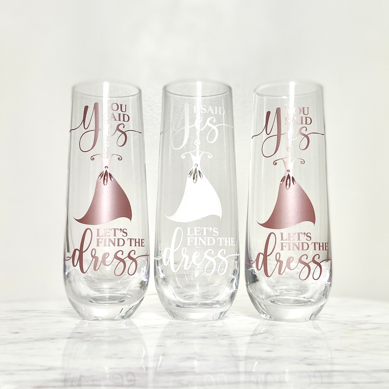 Pu&ograve; includere: Tre flute da champagne con la scritta "You said yes, let's find the dress" su di esse. Le flute sono in vetro trasparente con un design color oro rosa.