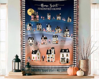 May include: A woven throw blanket featuring a gradient background from dark blue to orange, displaying the text "Home Sweet HAUNTED HOME" above a village of illustrated houses, ghosts, and figures, framed by a black and white checkered border.