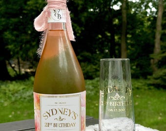 May include: A pink and gold bottle of sparkling wine with a personalized label that says "Sydney's 21st Birthday Aug 12, 2001". A clear champagne flute with a gold inscription that says "Sydney's 21st Birthday 08.12.2001" is next to the bottle.