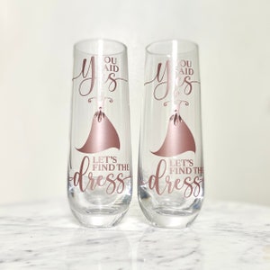 Pu&ograve; includere: Due flute da champagne con il testo "You said yes" e "Let's find the dress" in lettere dorate rosa. Le flute sono decorate con una silhouette di un vestito dorato rosa.