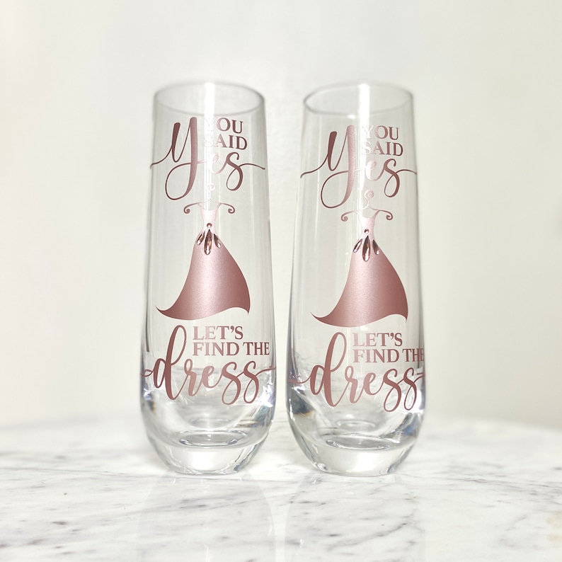 Pu&ograve; includere: Due flute da champagne con il testo "You said yes" e "Let's find the dress" in lettere dorate rosa. Le flute sono decorate con una silhouette di un vestito dorato rosa.