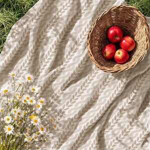 May include: A beige fabric blanket featuring a repeating white leaf pattern, a woven wicker basket containing four bright red apples, and a cluster of white daisies with yellow centers arranged on the fabric surface.
