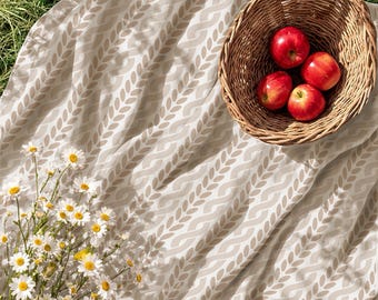 May include: A beige fabric blanket featuring a repeating white leaf pattern, a woven wicker basket containing four bright red apples, and a cluster of white daisies with yellow centers arranged on the fabric surface.