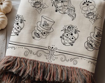 May include: A cream-colored woven throw blanket featuring a repeating black line art pattern of sugar skulls, some wearing top hats or floral crowns. The blanket has a horizontal decorative border and a multi-colored fringed edge at the bottom.