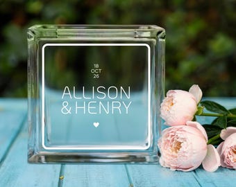May include: A clear glass block with the names "ALLISON & HENRY" and the date "18 OCT 26" printed in white. The block is on a blue wooden surface, with pink flowers in the foreground.