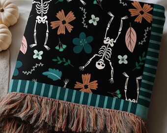 May include: A black throw blanket featuring a repeating pattern of white skeletons, orange flowers, teal blossoms, and green leaves. The blanket has a dark teal striped border and a multi-colored fringed edge with orange and grey threads.