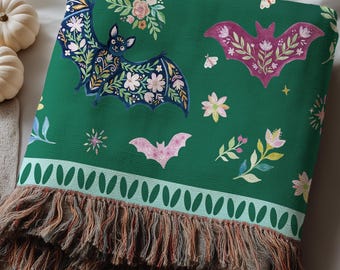 May include: A dark green woven throw blanket featuring a repeating pattern of bats with floral wings in shades of pink, blue, and white. The blanket includes a light green border with a leaf motif and a multi-colored fringed edge at the bottom.