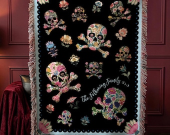 May include: A black woven throw blanket featuring a repeating pattern of floral-filled skulls and crossbones in various sizes. The blanket has a light blue border with a scalloped edge, fringed sides, and the text The Folkening Family in white script.